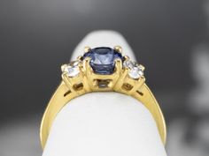 This timeless 18 karat yellow gold mounting is classic and simple and is set with a lovely deep navy blue sapphire, accented on each side by bright high-quality diamonds. The warmth of the yellow gold is an amazing contrast with the beautiful color of the sapphire, which twinkles with a dark and mysterious blue that pops hints of indigo and teal when moved in the light. Metal: 18K Yellow Gold Gem: Sapphire 1.73 Carats Gem Measurements: 7.3 x 5.8 mm, Oval Accents: 2 Diamonds totaling .53 Carats, Gia Certified Gold Sapphire Ring, Sapphire And Diamond Engagement Ring, Dark And Mysterious, Deep Navy Blue, Quality Diamonds, Eternity Bands, Diamond Engagement Ring, Sapphire Ring, Blue Sapphire