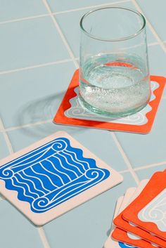 three coasters with blue and orange designs on them sitting on a tile floor next to a glass of water