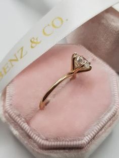 a gold ring with a diamond on it in a pink velvet case, next to a white ribbon