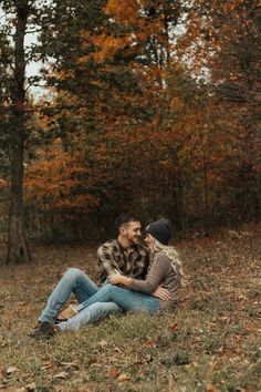 a man and woman sitting on the ground in front of trees with autumn leaves around them