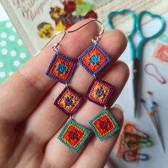 a hand holding four square shaped crocheted earrings in front of scissors and other crafting supplies