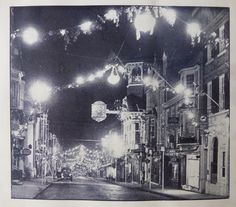 an old black and white photo of a street at night with lights strung from the buildings