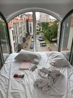 an unmade bed in front of a large window with cars outside it and books on the bed