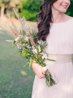 a woman in a white dress holding a bouquet of flowers and greenery smiling at the camera