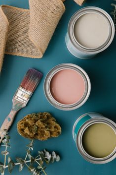 three cans of paint next to some flowers and a brush on a blue surface with burlap