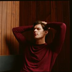 a man in a red shirt leaning against a wooden wall with his hands on his head