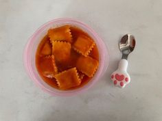 a plastic bowl filled with ravioli on top of a table next to a spoon