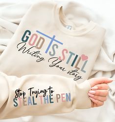 someone holding up a sweatshirt that says god is still when you say so stop trying to steal the pen