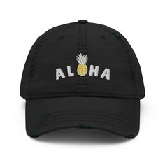 A tropical blend of laid-back style and island vibes, perfect for those who carry a bit of paradise wherever they go! Crafted from durable, distressed black fabric, this cap exudes effortless cool with a touch of beachy charm. The front of the cap features the cheerful phrase "Aloha" embroidered in vibrant colors, accompanied by a cute pineapple, symbolizing hospitality and tropical fun. It's a whimsical nod to the spirit of Hawaii, bringing sunshine and smiles no matter where you are. With its classic baseball cap design, distressed fabric, and adjustable strap, this cap offers a comfortable and customizable fit for island dreamers of all ages. Whether you're lounging by the pool, exploring the city, or daydreaming of sandy shores, this cap is your perfect companion for adding a splash of Distressed Black Hat For Summer, Casual Cotton Trucker Hat For Vacation, Black Distressed Hat For Summer, Adjustable Dad Hat For Vacation, Adjustable Dad Cap For Vacation, Casual Baseball Cap For Beach Season Vacation, Summer Adjustable Dad Hat For Vacation, Casual Adjustable Trucker Hat For Vacation, Summer Dad Hat For Beach