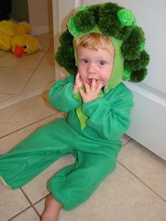 a little boy in a green costume sitting on the floor with his hands under his mouth