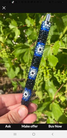 a hand holding a blue and white beaded pen in front of some green plants