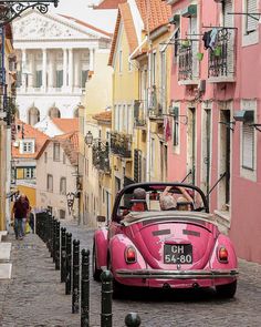 a pink car driving down a cobblestone road in front of tall colorful buildings