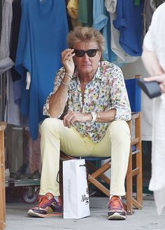 an older man sitting on a chair talking on his cell phone while holding a shopping bag