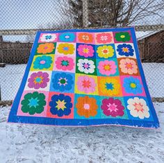 a colorful quilt sitting on top of snow covered ground next to a chain link fence