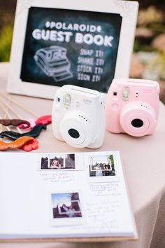 a table topped with pink and white cameras next to a chalkboard sign that says polaroid guest book