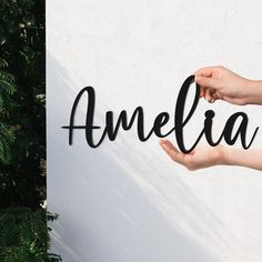 two hands are holding the word amelie against a white wall with green leaves in front of it