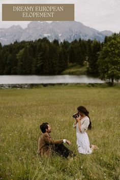 a man and woman sitting on the grass in front of a lake with mountains behind them