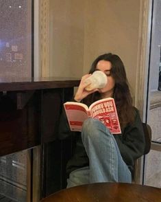 a woman sitting in a chair drinking from a coffee cup and reading a book with her legs crossed