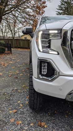 the front end of a white truck parked on gravel with trees in the back ground