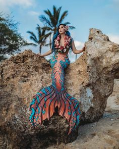 a woman in a blue and red mermaid costume standing on rocks with palm trees behind her