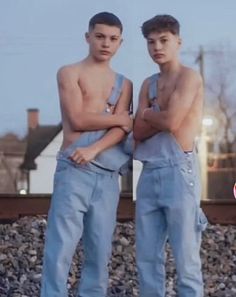 two young men standing next to each other in front of a train track with their arms crossed