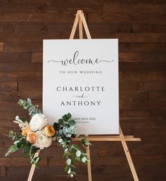 a welcome sign with flowers and greenery is displayed on an easel in front of a wooden wall