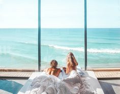 two people sitting on a bed looking out at the ocean