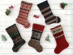 four knitted christmas stockings laid out on a white wooden surface with holly and pine cones