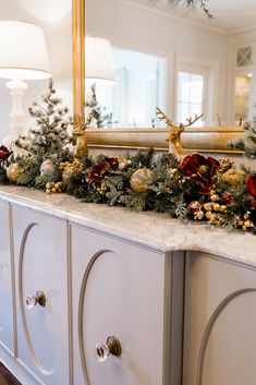 a white dresser topped with christmas decorations and greenery next to a gold framed mirror