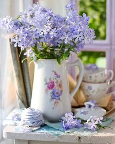 purple flowers are in a white pitcher on a window sill next to teacups