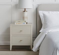 a nightstand with two books on top of it next to a white bed and lamp