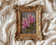 a painting of pink flowers in a gold frame on a white bed linens covered sheet