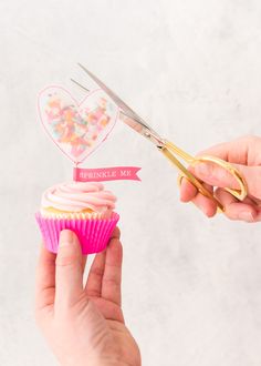 someone is holding a cupcake with pink frosting and a pair of gold scissors