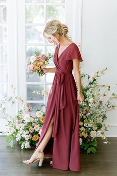 a woman standing in front of flowers wearing a long red dress and holding a bouquet