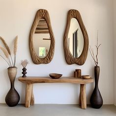 two mirrors are hanging on the wall above a wooden table and vases with dried flowers