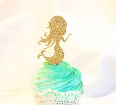 there is a cupcake with blue and green frosting on it that has a gold glitter mermaid figure on top