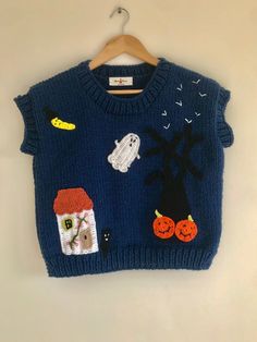 a blue sweater with pumpkins, ghost and house on the front is hanging from a wooden hanger