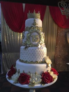 a white and gold wedding cake with red roses on the bottom, surrounded by curtains