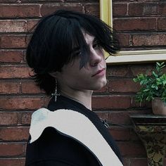 a man with black hair and piercings standing in front of a brick wall next to a potted plant