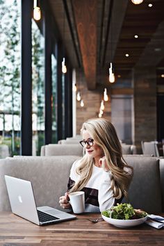 a woman sitting at a table in front of a laptop computer and holding a coffee cup