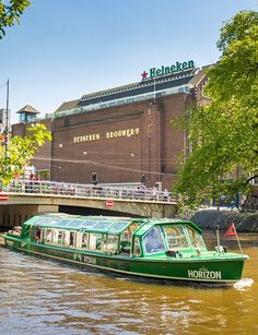 a green boat traveling down a river next to a tall brick building with a sign that reads heineken