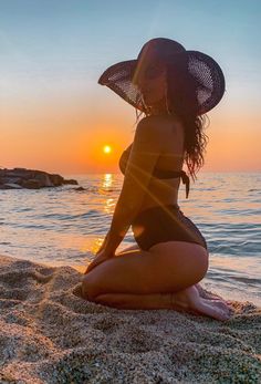 a woman sitting on top of a sandy beach next to the ocean wearing a hat