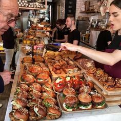 a man and woman standing in front of a counter filled with sandwiches, burgers and other foods