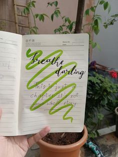 someone holding up a book with writing on it in front of a potted plant