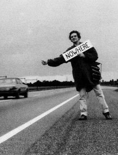 a man holding a sign that says nowhere on the side of a road next to a car