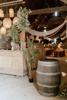 This shows a wedding bar display in a rustic barn. The bar display features fake trees, wooden crates and vases of dried flowers. Next to the bar is a rustic wooden barrel. Above the bar are cane lampshades with warm light bulbs. Wedding Buffet, Bar Set Up