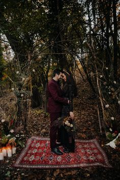 a man and woman standing on top of a rug in the woods surrounded by candles