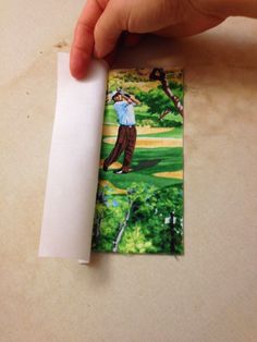 a hand holding a piece of paper with a golf scene on it and another person taking a photo