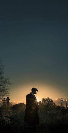 a man in a long coat and hat standing in the grass near graves at sunset