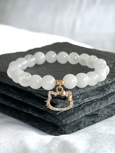 White Jade is said to be a calming and soothing stone for the mind and body. It is believed to promote feelings of abundance, comfort, and peacefulness. Many today use White Jade to connect us to higher vibrational energies and stay focused on our end goals. DETAILS ------------ - Genuine White Jade beads, 8mm - 18k gold plated spacers, 6mm - 18k gold plated cubic zirconia kitty charm, 15mm x 10mm - Double-corded with durable and flexible elastic cord  - Meaning card and polishing cloth included Adjustable White Jewelry With Cat Design, Adjustable White Cat Design Jewelry, Cat With Bow, Y2k Cat, Button Pouch, Bracelet Y2k, Hello Cat, Cat Charm Bracelet, Kawaii Kitty